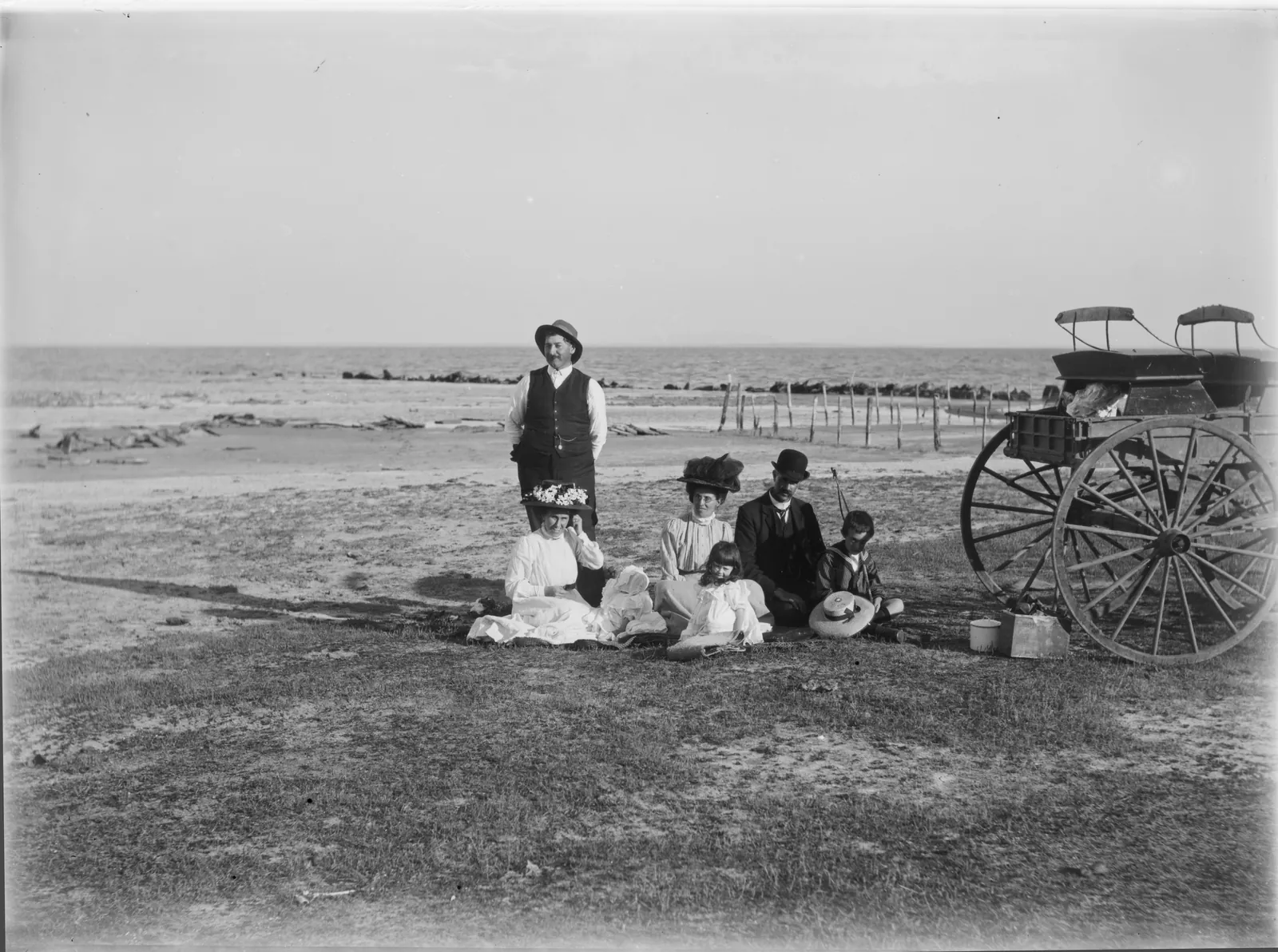 A family picnicking on a grassy shore, a man standing behind women and children seated on a rug, a two-wheeled buggy with its shafts down beside them, and a flat lake running to the horizon behind.