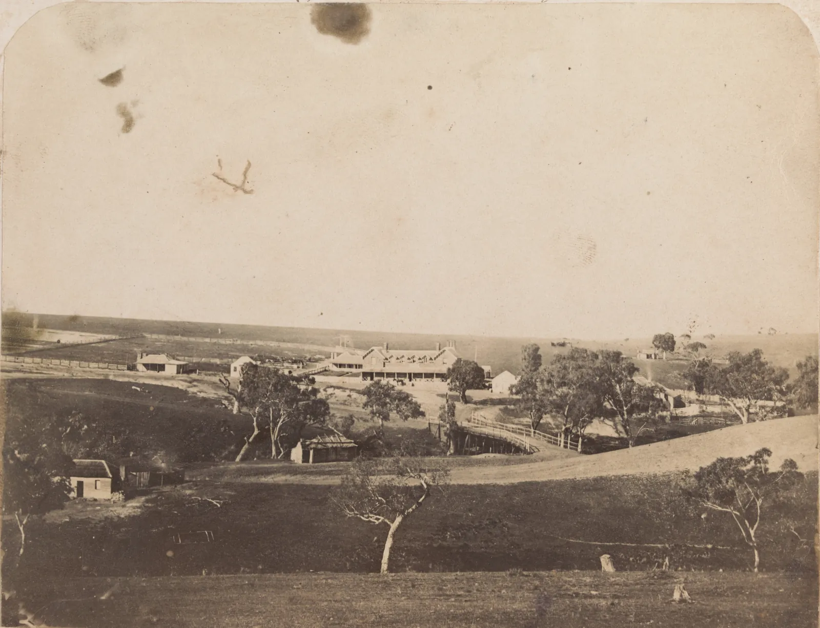 An elevated view across a grassy valley: a timber road bridge with a post-and-rail handrail crosses the creek, the road curving up to a long two-storey hotel with a verandah, sheds and cottages around it, a slab hut among gum trees in front, and the open plain running to the horizon.